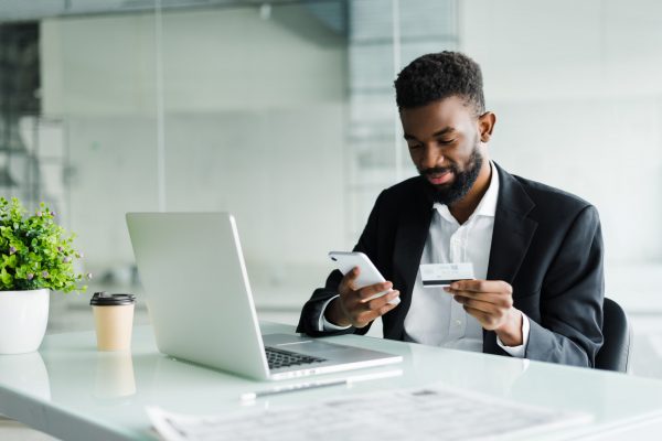 African American man in casual yellow tshirt paying with credit card online while making orders via mobile Internet. Attractive black hipster male making transaction using mobile bank application.